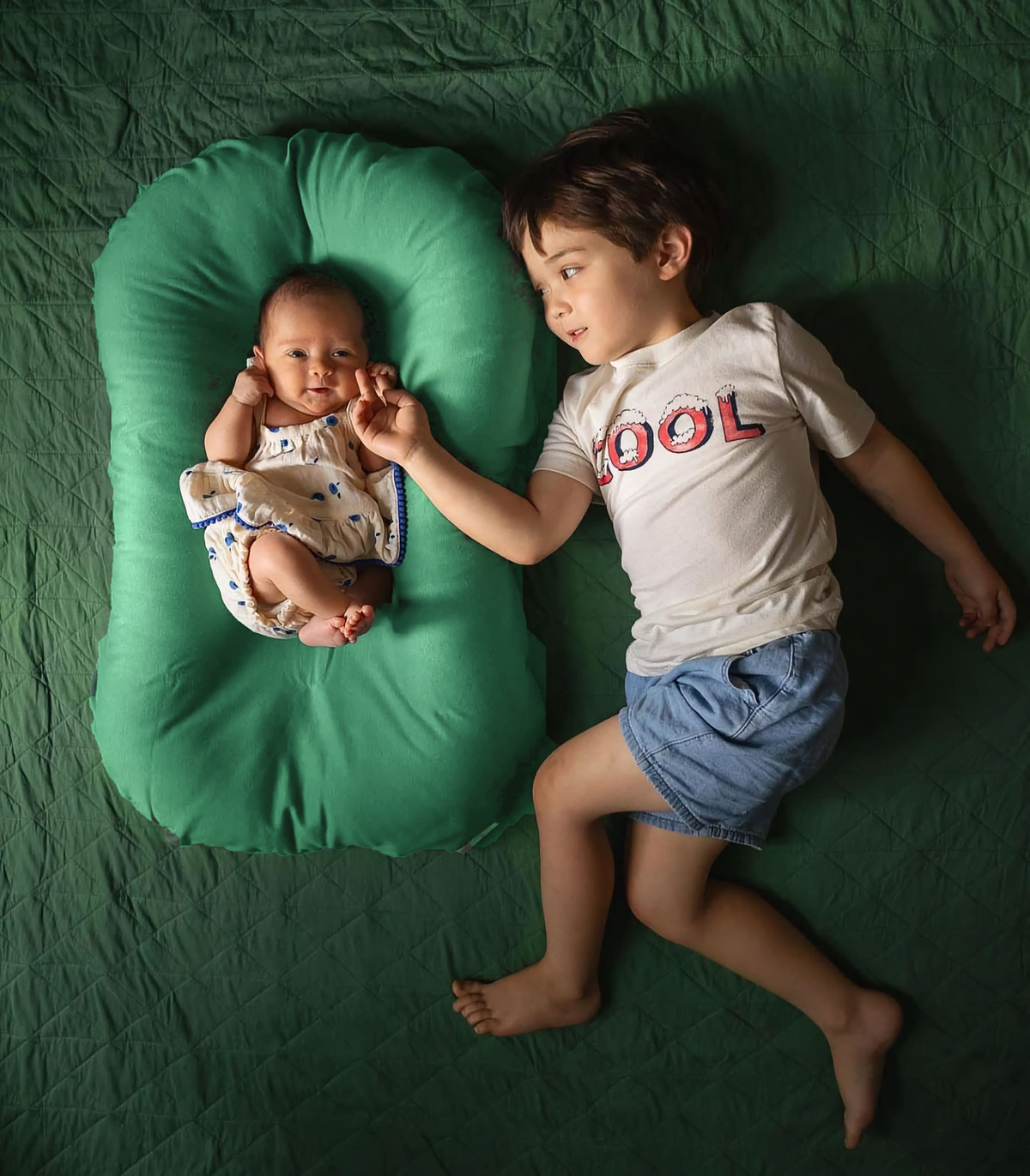 Toddler lying next to a baby in a Moss green infant lounger, overhead view